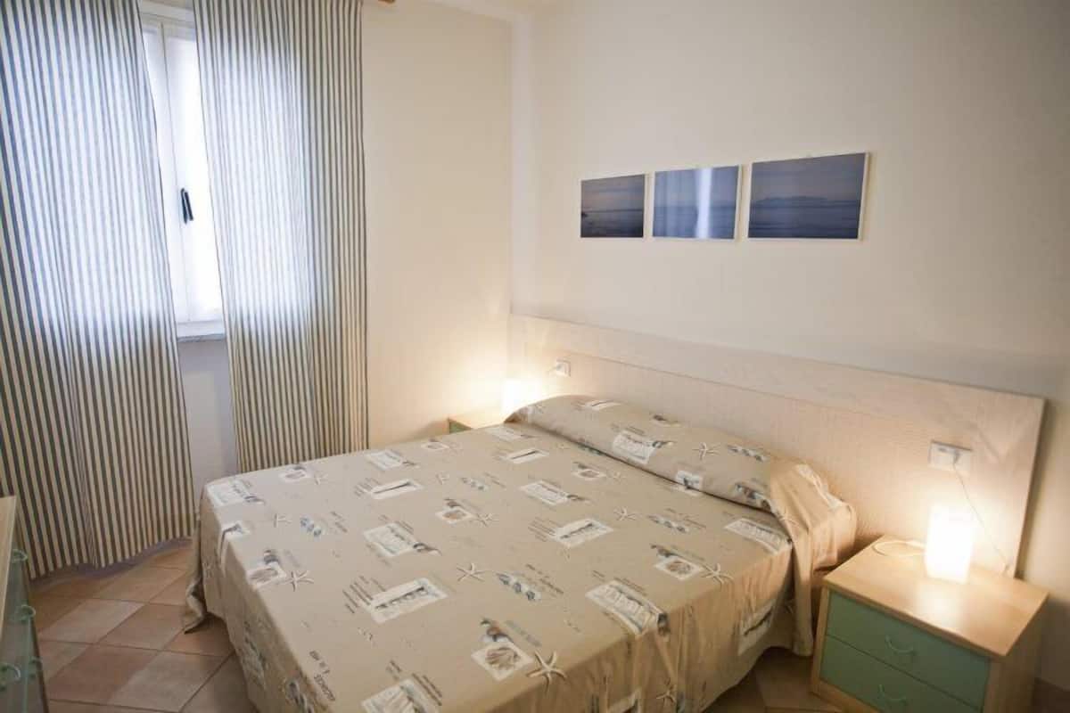 A bedroom with a bed, a nightstand, a drawer, and a window with curtains. There are three framed pictures on the wall.