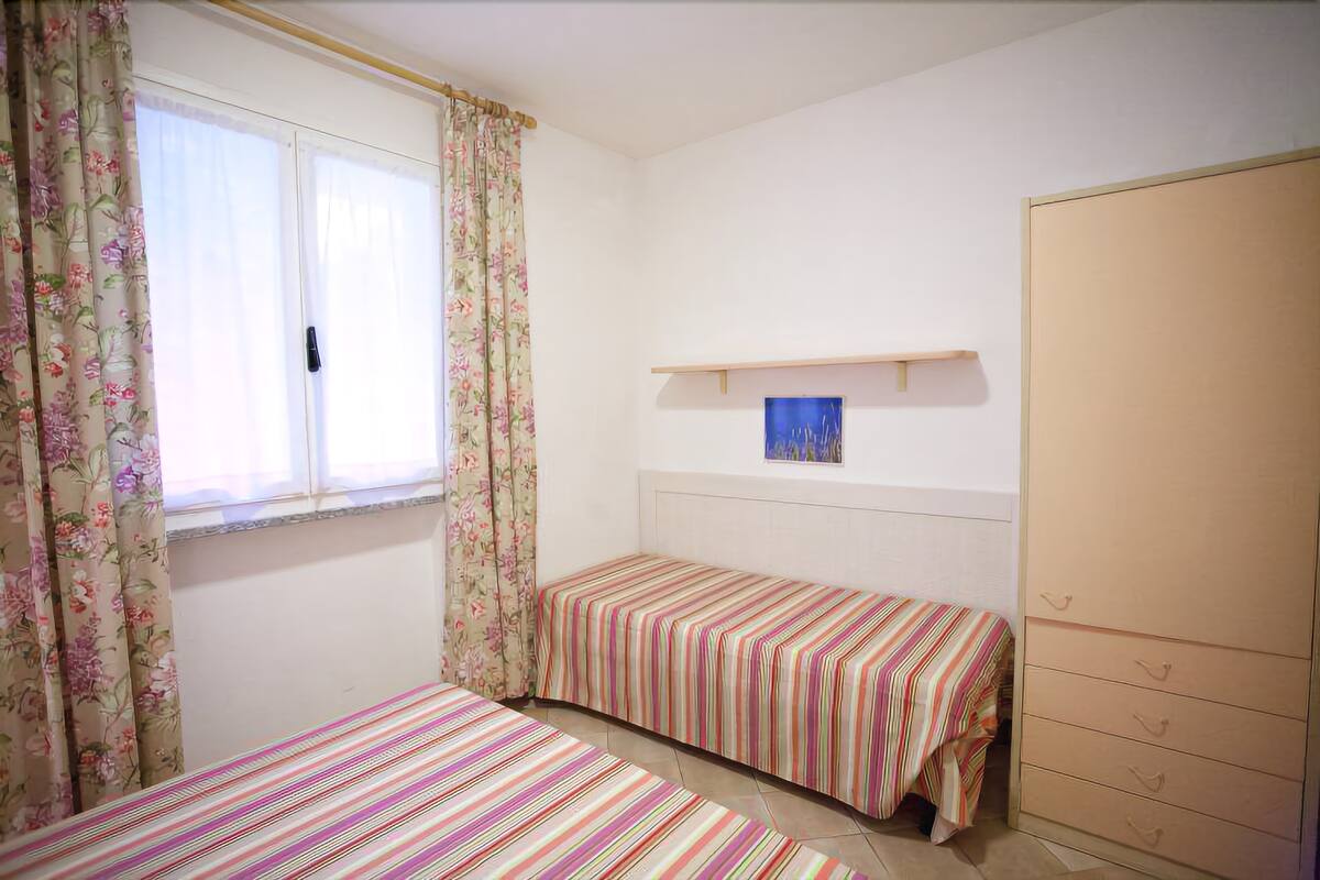 A bedroom with two single beds, a wardrobe, a window with floral curtains, and a small shelf with a painting.