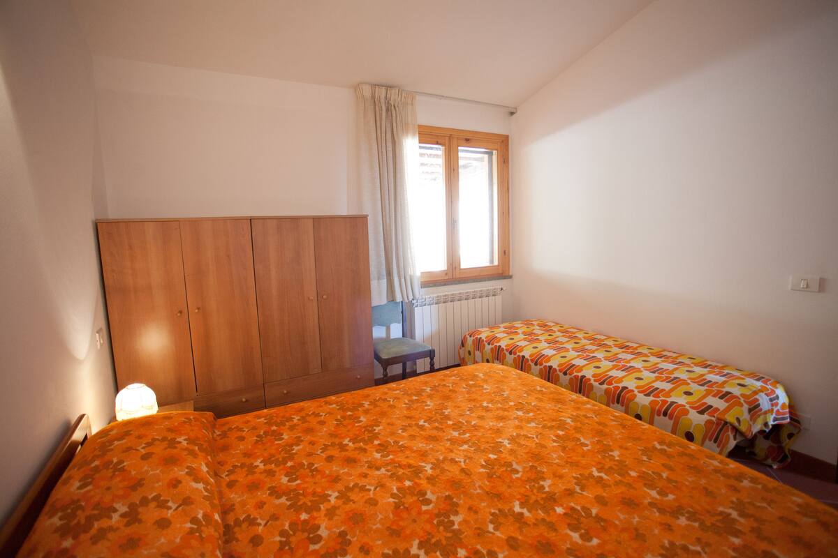 A bedroom with two beds, a wooden wardrobe, a chair, and a window with curtains.