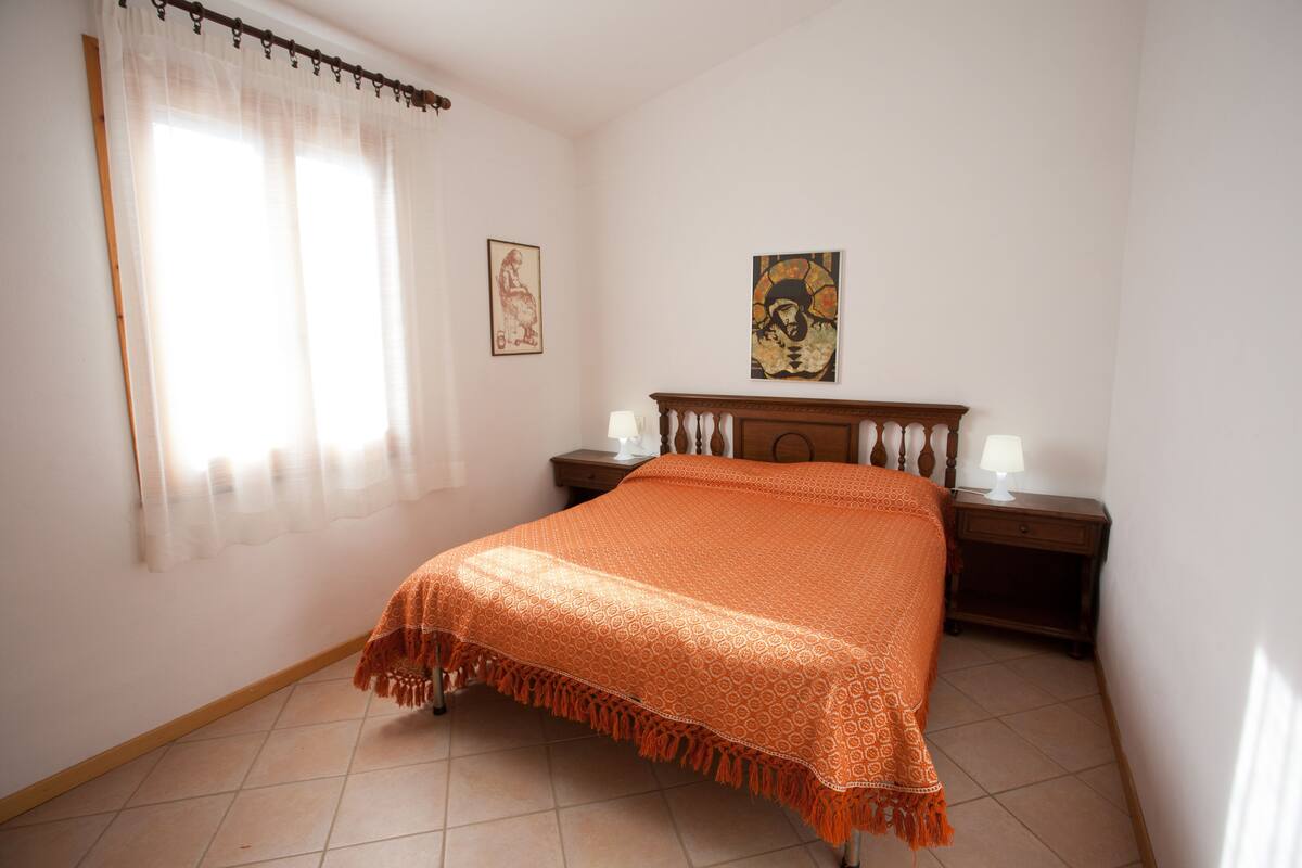 A bedroom with a bed, a nightstand, a lamp, a window with sheer curtains, and two wall-mounted artworks.