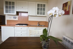 Cocina con gabinetes blancos, encimera de madera, estufa y fregadero. Hay una silla blanca y una mesa con una orquídea en maceta.