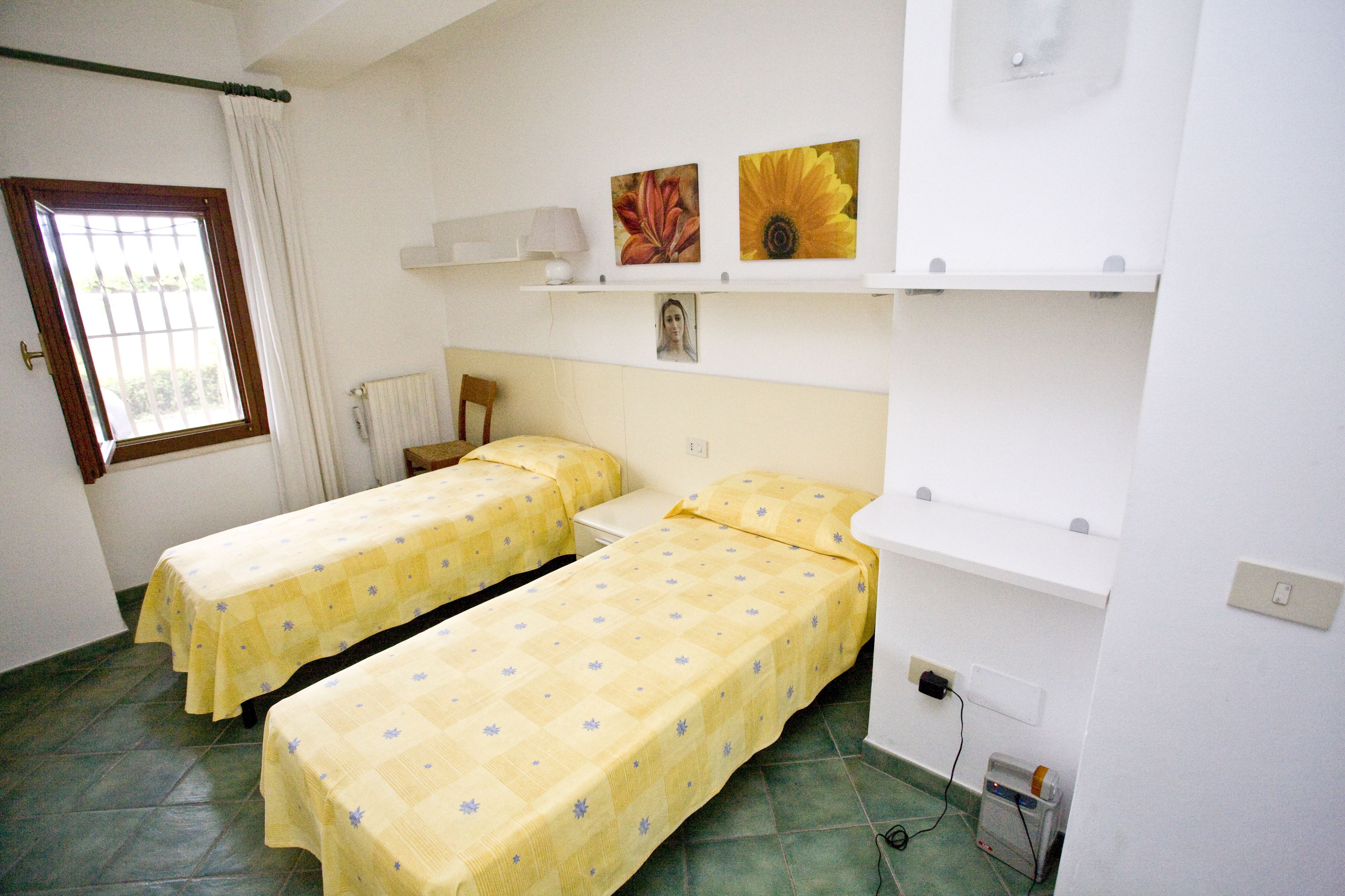 A room with two single beds, a window with curtains, a radiator, and a shelf with pictures.