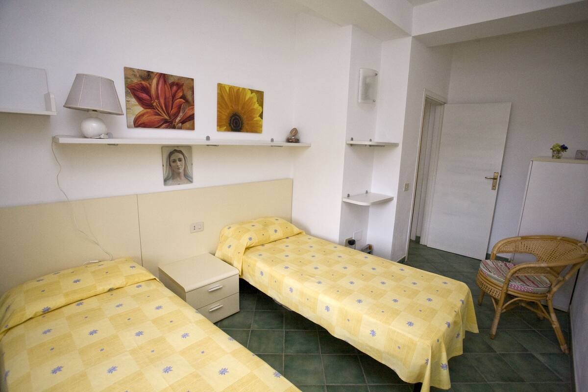 Una habitación pequeña y limpia con dos camas individuales, una silla, una mesita de noche y un estante en la pared con adornos.