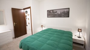 A bedroom with a green bedspread, a cityscape picture on the wall, a bedside table with a lamp, and a door leading to another room. - Appartamento Bilo Amore (Portoferraio)