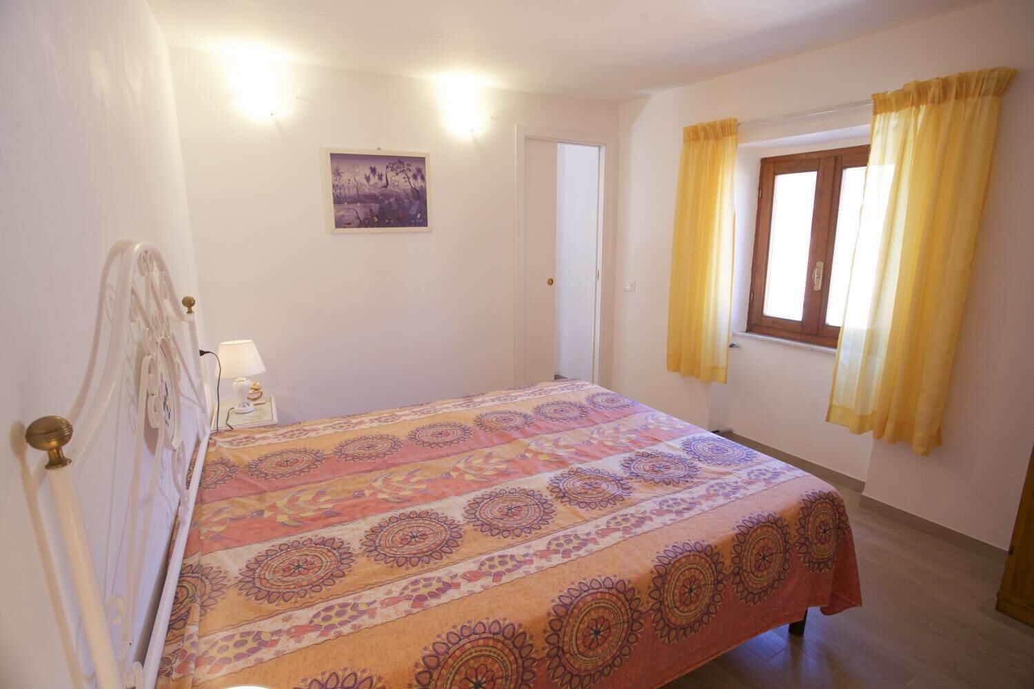 Une chambre à coucher avec un lit, une table de chevet, une lampe fixée au mur, une porte et une fenêtre avec des rideaux jaunes.
