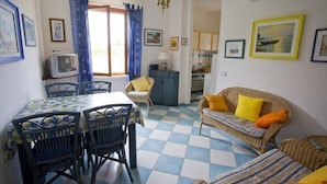 A room with a checkered floor, a dining table with chairs, a sofa with cushions, a television, and a kitchen area. - Appartamento Casa Paoletta (Rio)