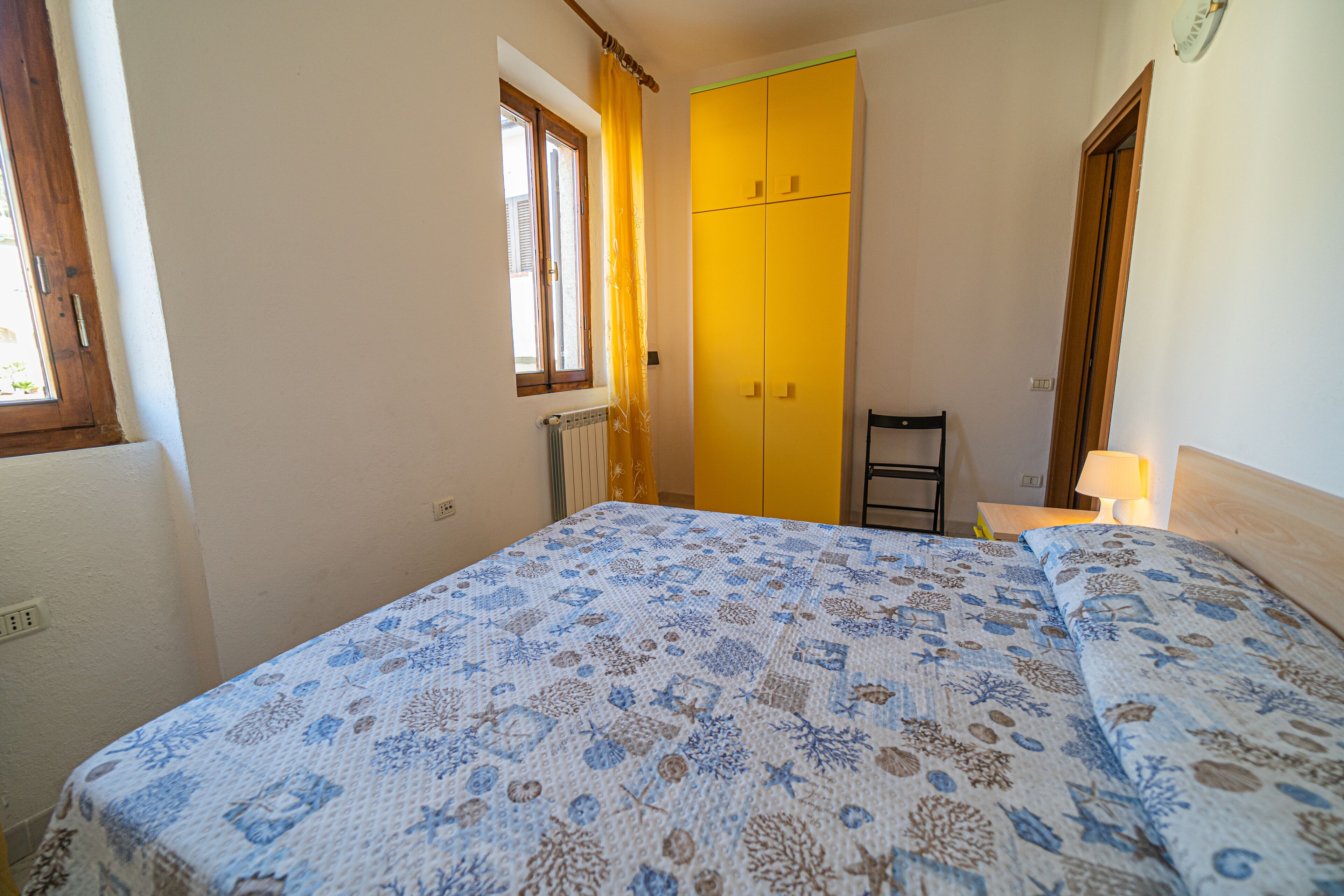 A bedroom with a bed, a window, a radiator, a yellow wardrobe, and a nightstand with a lamp.