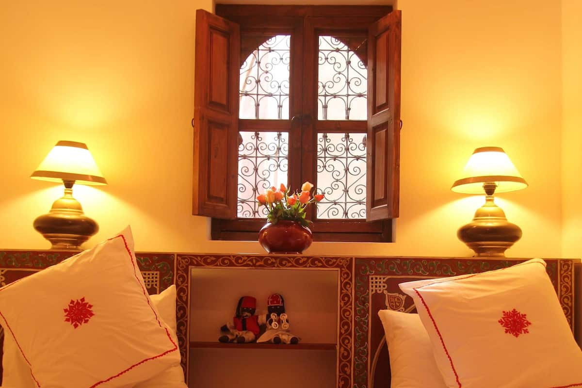 A room with two beds, each with a checkered bedspread and a red floral pillow. There are two wall-mounted lamps on either side of the window, which has a decorative iron grill. A small shelf with figurines is visible under the window.