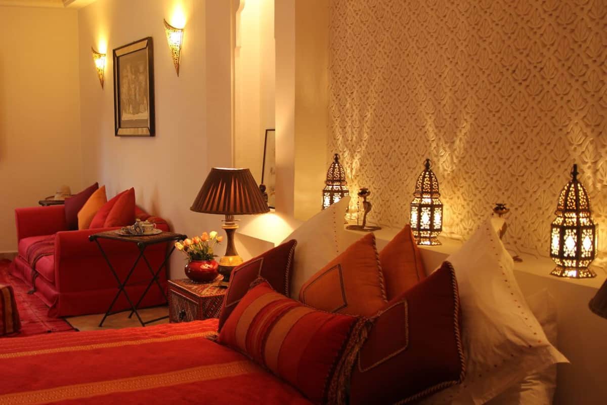 A room with a bed, red sofa, and ottomans. There are wall sconces and a decorative lamp.