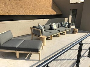 A sunlit outdoor seating area with a thatched roof, cushioned lounge chairs, and a wooden table. - Bakubung Self-Catering Chalets (Pilanesberg National Park)