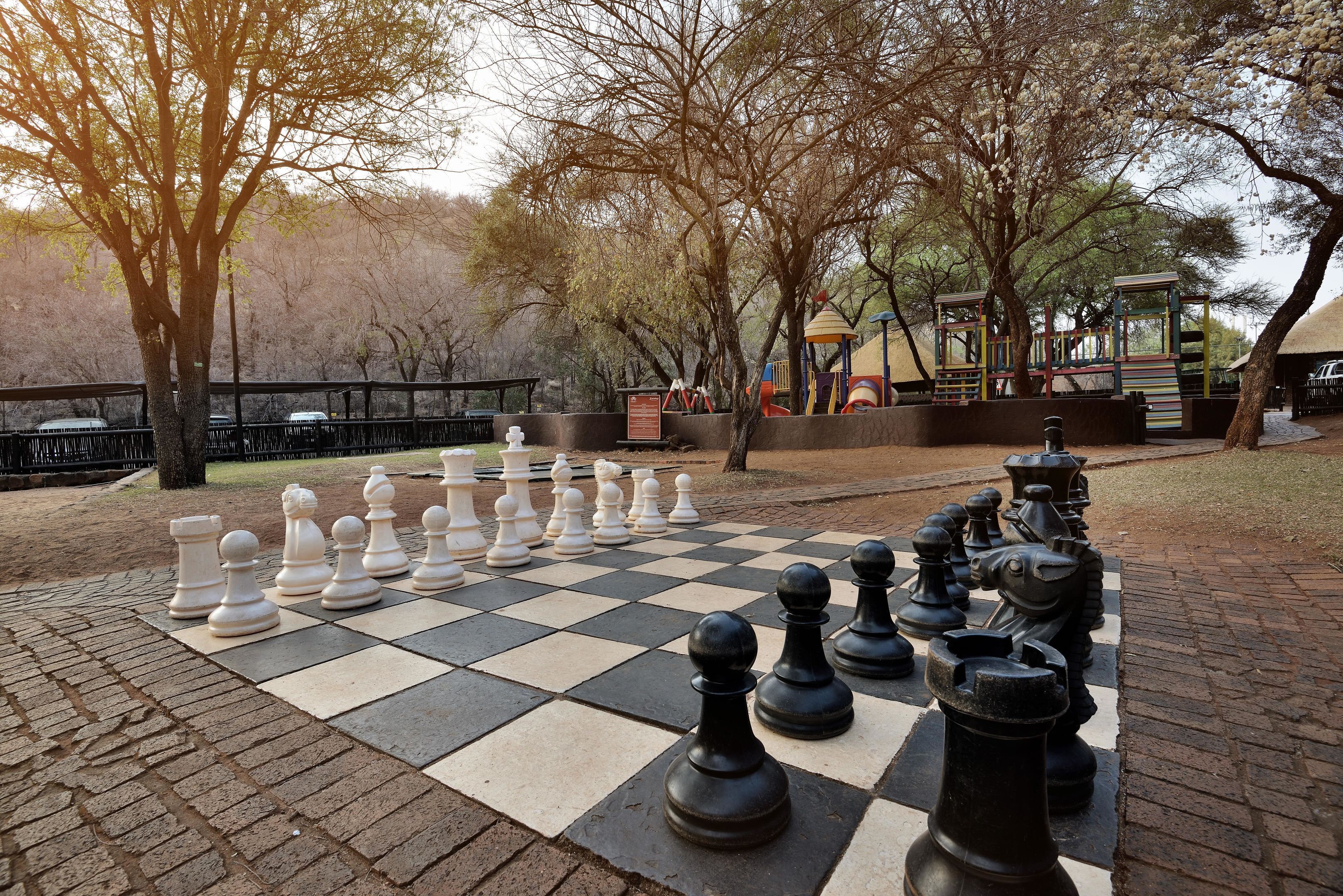 Un tablero de ajedrez grande al aire libre en un parque, con árboles y juegos infantiles al fondo.