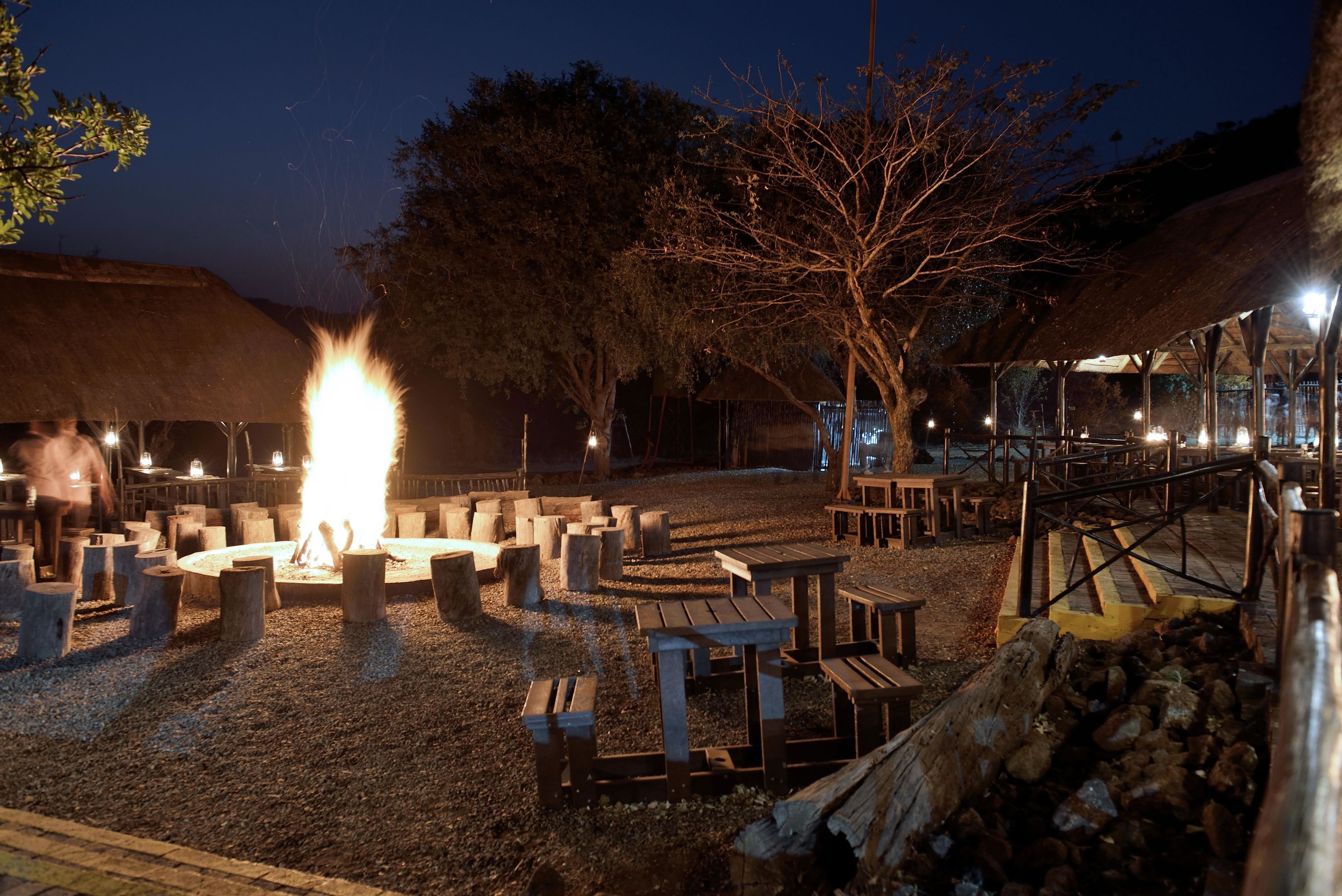 An outdoor seating area with a central fire pit, surrounded by wooden benches and tables, set against a backdrop of trees and a thatched-roof building.