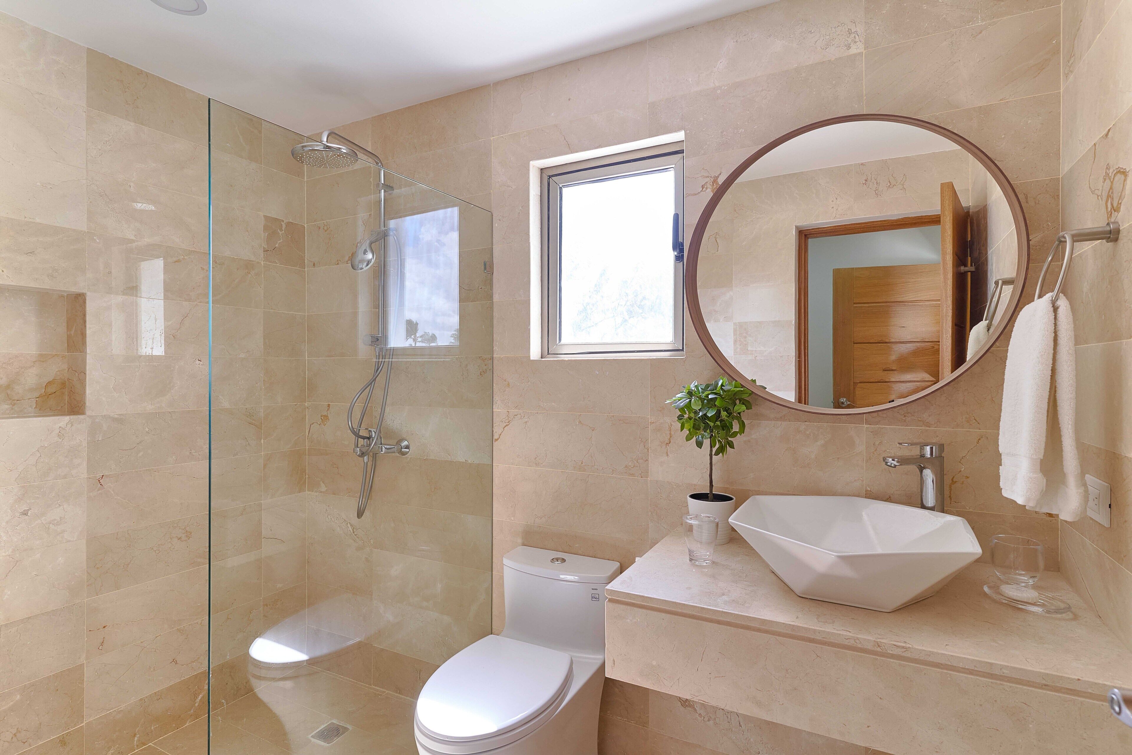 A modern bathroom with a glass-enclosed shower, a white toilet, a sink with a large vessel basin, and a round mirror.