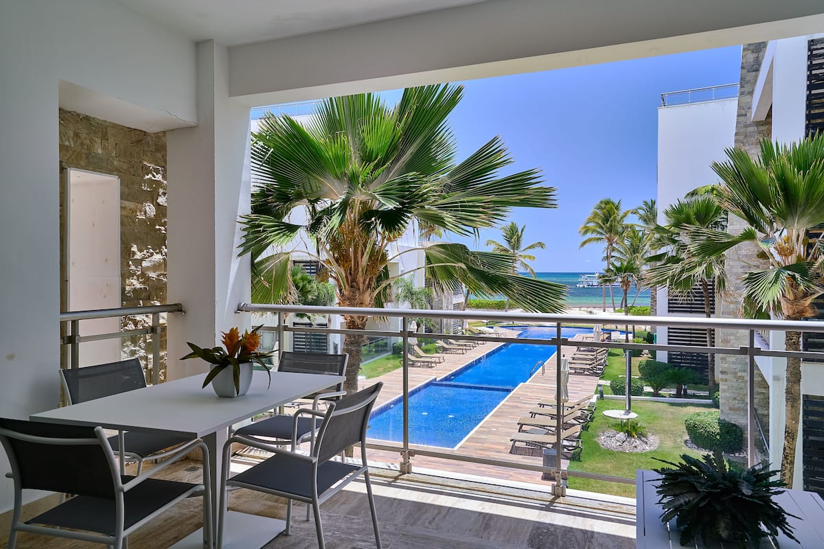 A balcony with a view of a swimming pool, palm trees, and a clear blue sky.