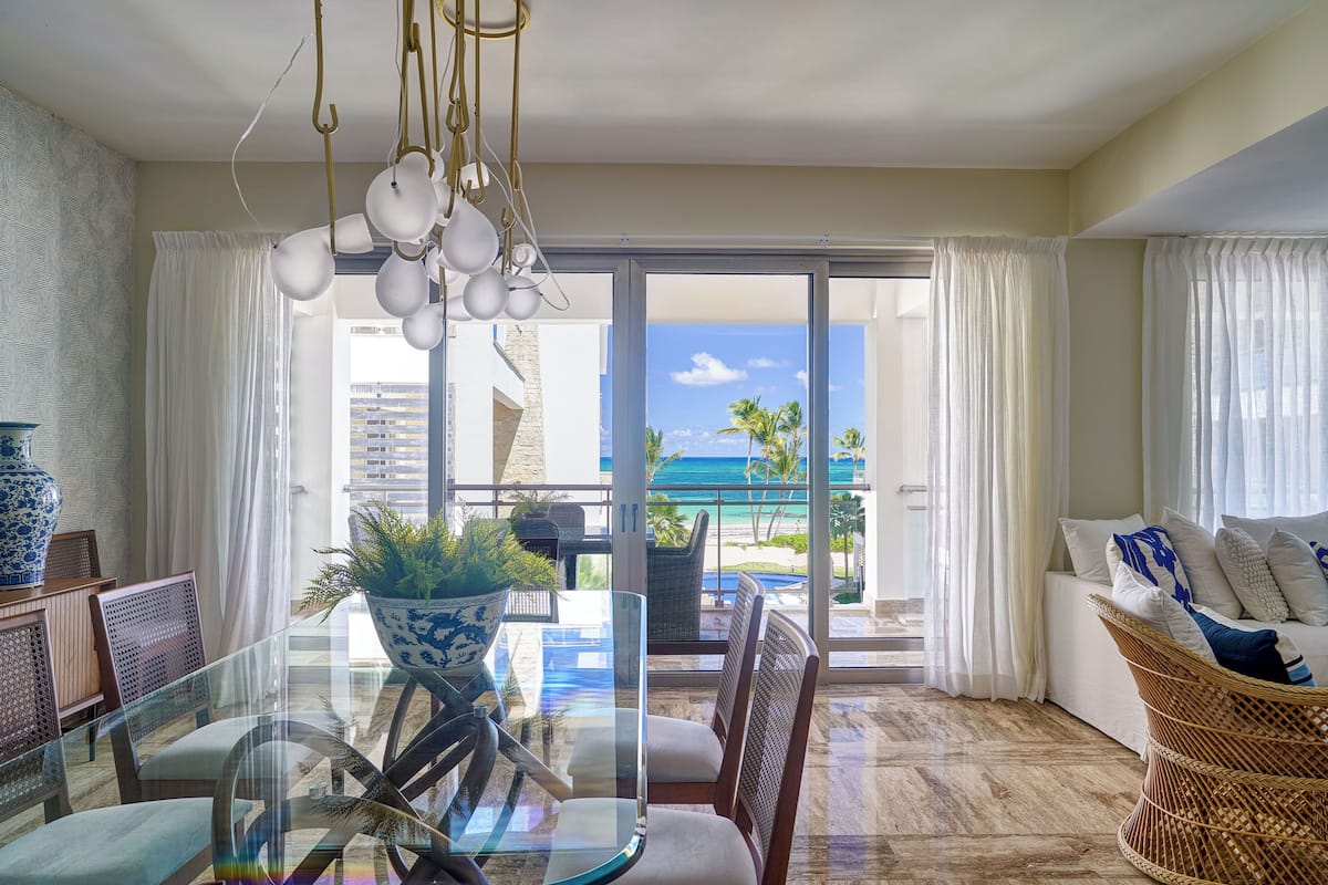 A modern living room with a glass table, wicker chairs, a sofa with cushions, a potted plant, and a view of the ocean.