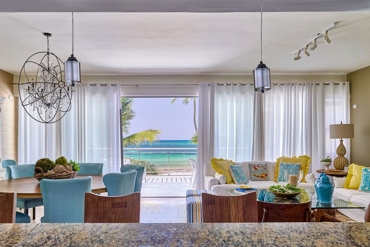 A spacious living area with a view of the ocean, a dining area with blue chairs, and a white sofa with yellow cushions.