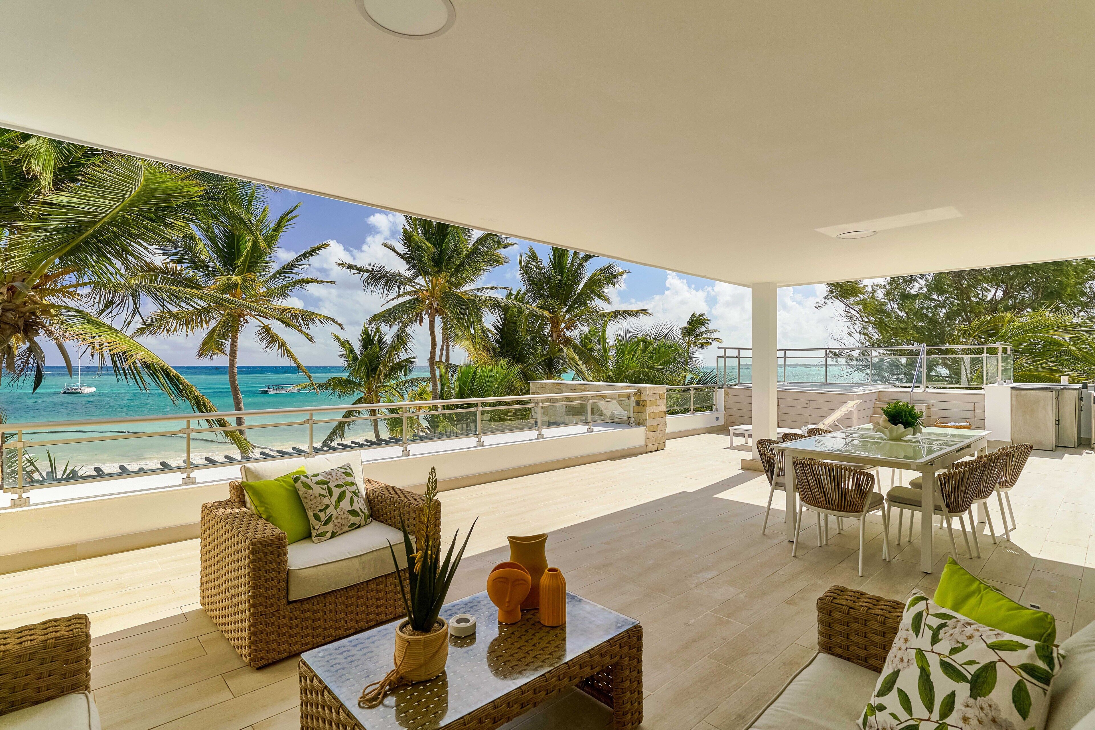 Ein geräumiger Balkon mit Blick auf das Meer und Palmen, möbliert mit Korbmöbeln und einem Glastisch.