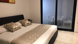 Un dormitorio con cama, almohadas, una mesita de noche y vistas a sillas exteriores a través de un ventanal.