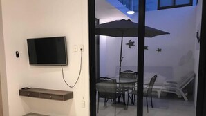 Una sala moderna con un televisor de pantalla plana montado en la pared, un vidrio que da a un comedor con una mesa y sillas, y una lámpara en el techo.