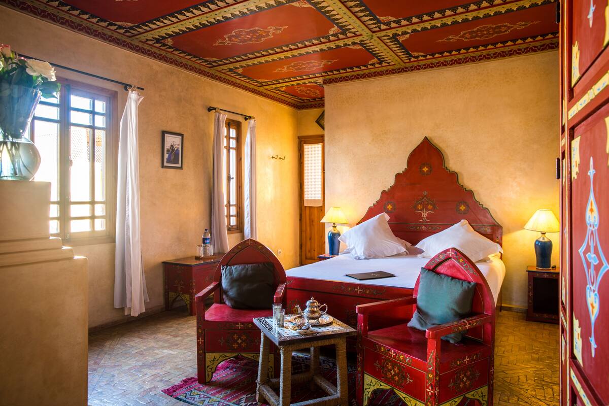 Una habitación tradicional con cama, dos sillas, una mesita y un techo decorativo.