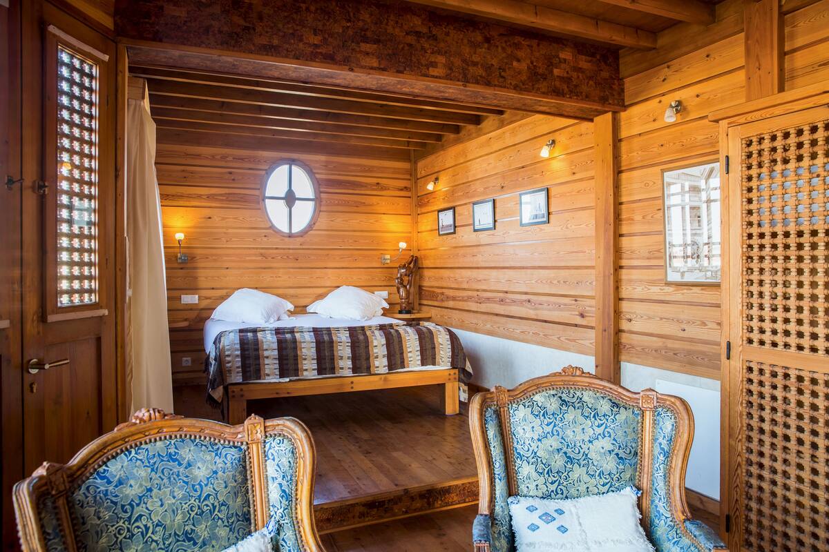 Un dormitorio de cabaña de madera con una cama, dos sillas y una ventana con diseño de celosía de madera.