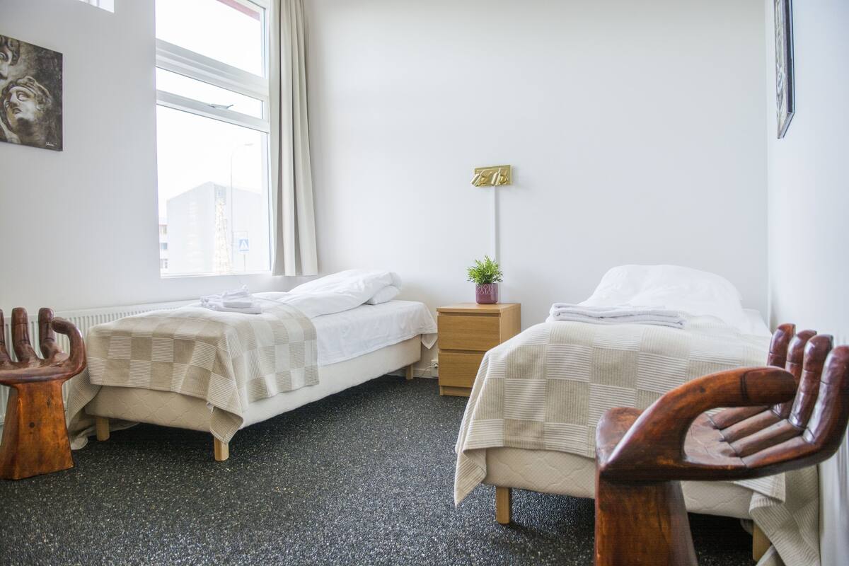 A hotel room with two beds, a small wooden nightstand, a potted plant, and a window with a view of buildings.