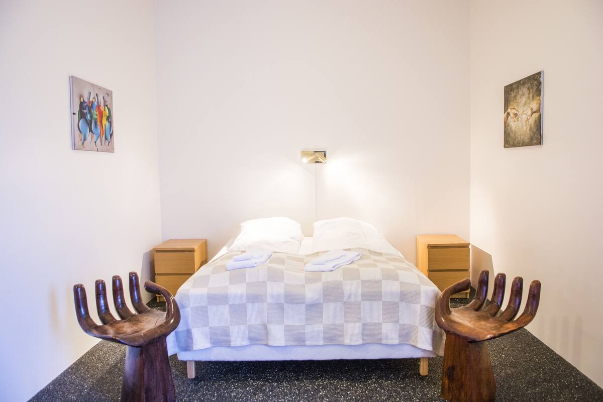 A bedroom with a bed, two nightstands, a wall-mounted light, and two wooden hand-shaped stools.