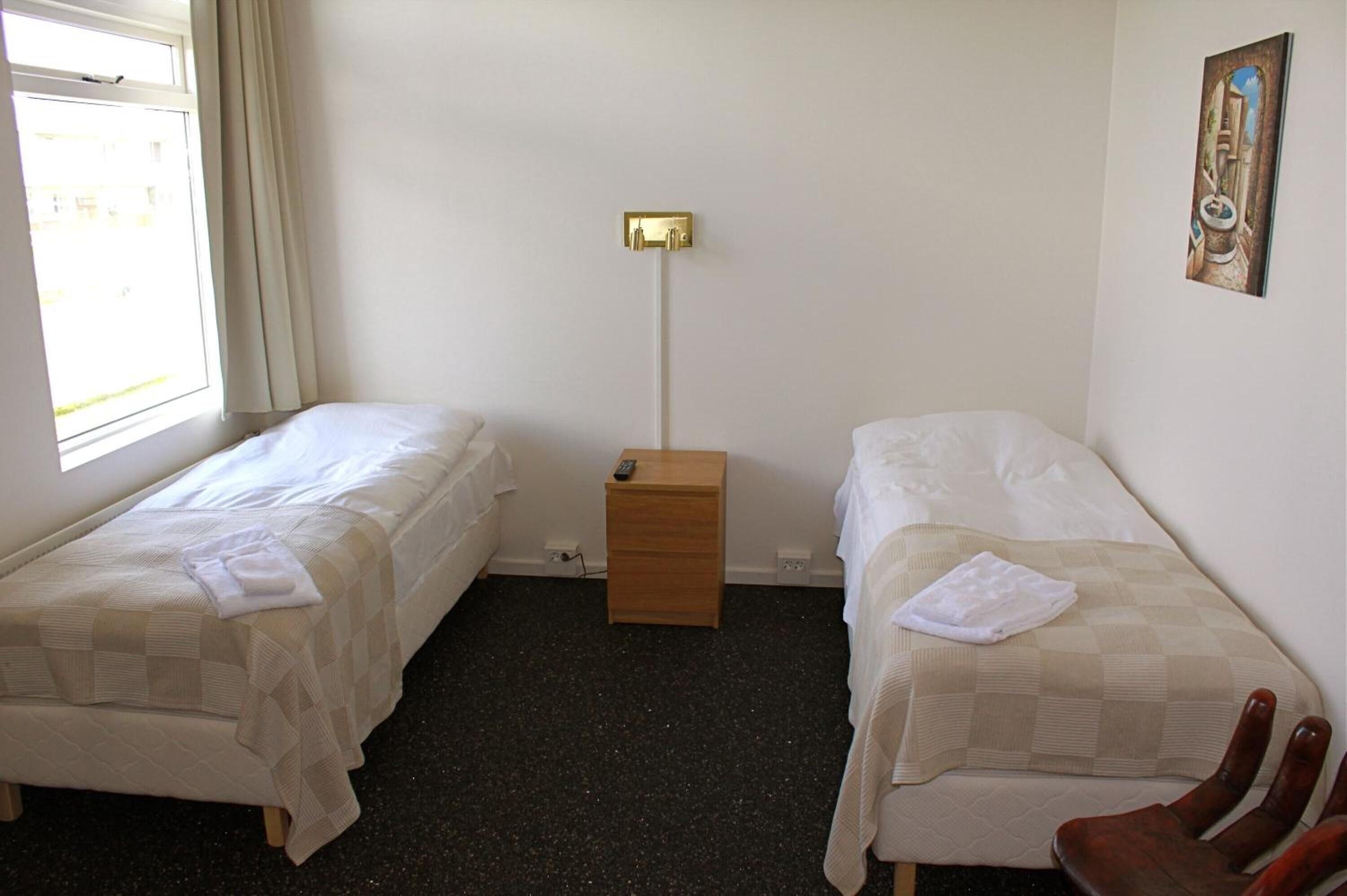 A room with two single beds, a bedside table with a lamp, a chair, and a window with curtains.