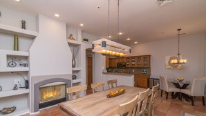 A spacious dining area with a wooden table and chairs, a fireplace, and a kitchen with wooden cabinets and a hanging light fixture. - Casa Delight (Scottsdale)