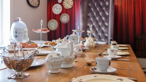 Uma mesa de jantar posta com diversos utensílios de chá e doces, com um relógio e cortinas ao fundo.