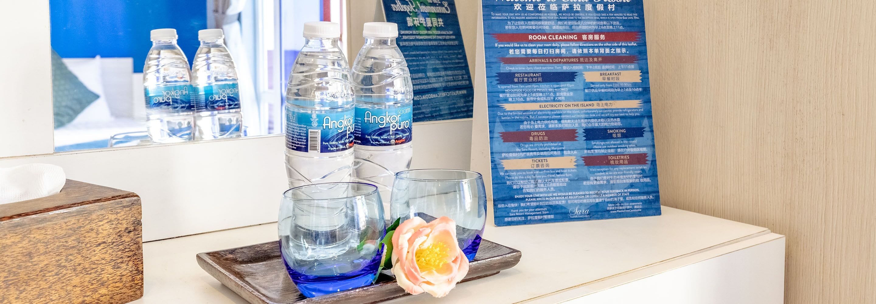 A hotel reception area with a glass display of water bottles, a tray with glasses and a rose, and a multilingual informational board.