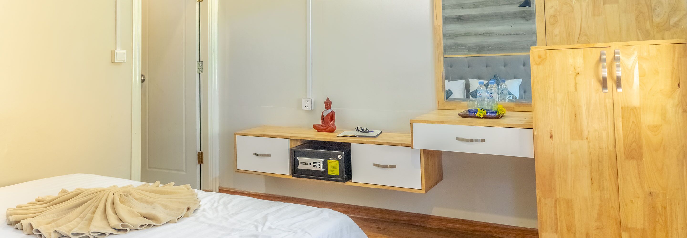 A hotel room with a bed, a wooden wardrobe, a desk with drawers, a mirror, and a small statue.