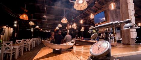 A rustic interior with wooden tables and chairs, hanging pendant lights, and a vintage clock on the counter.