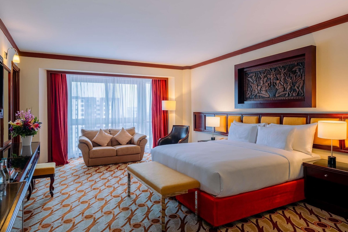 Ein Hotelzimmer mit einem großen Bett, einem Schlafsofa, einer Bank, einem Schreibtisch und einem Fenster mit Vorhängen.