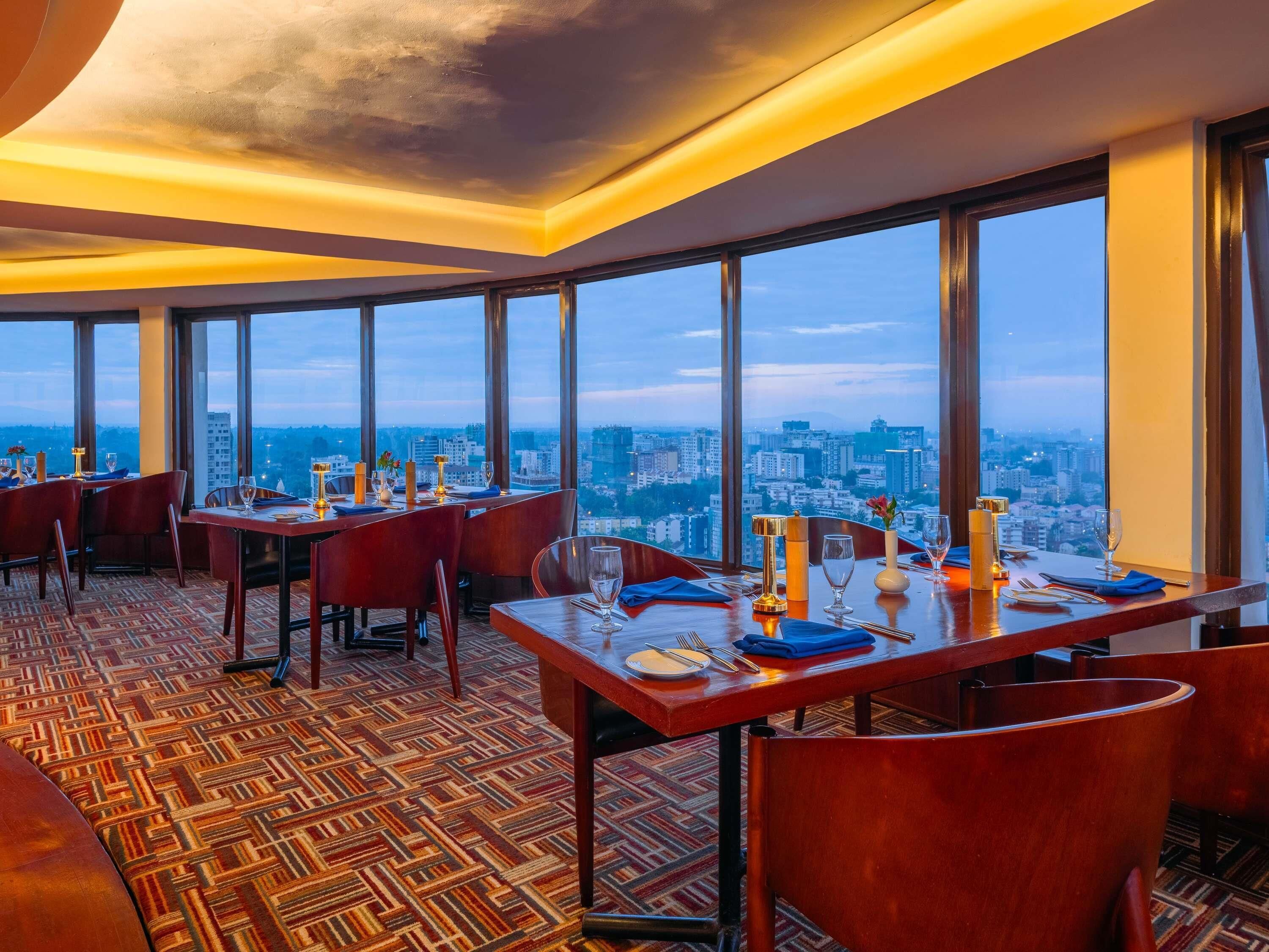 A high-rise restaurant with multiple tables set for dining, large windows offering a city view, and a patterned carpet.