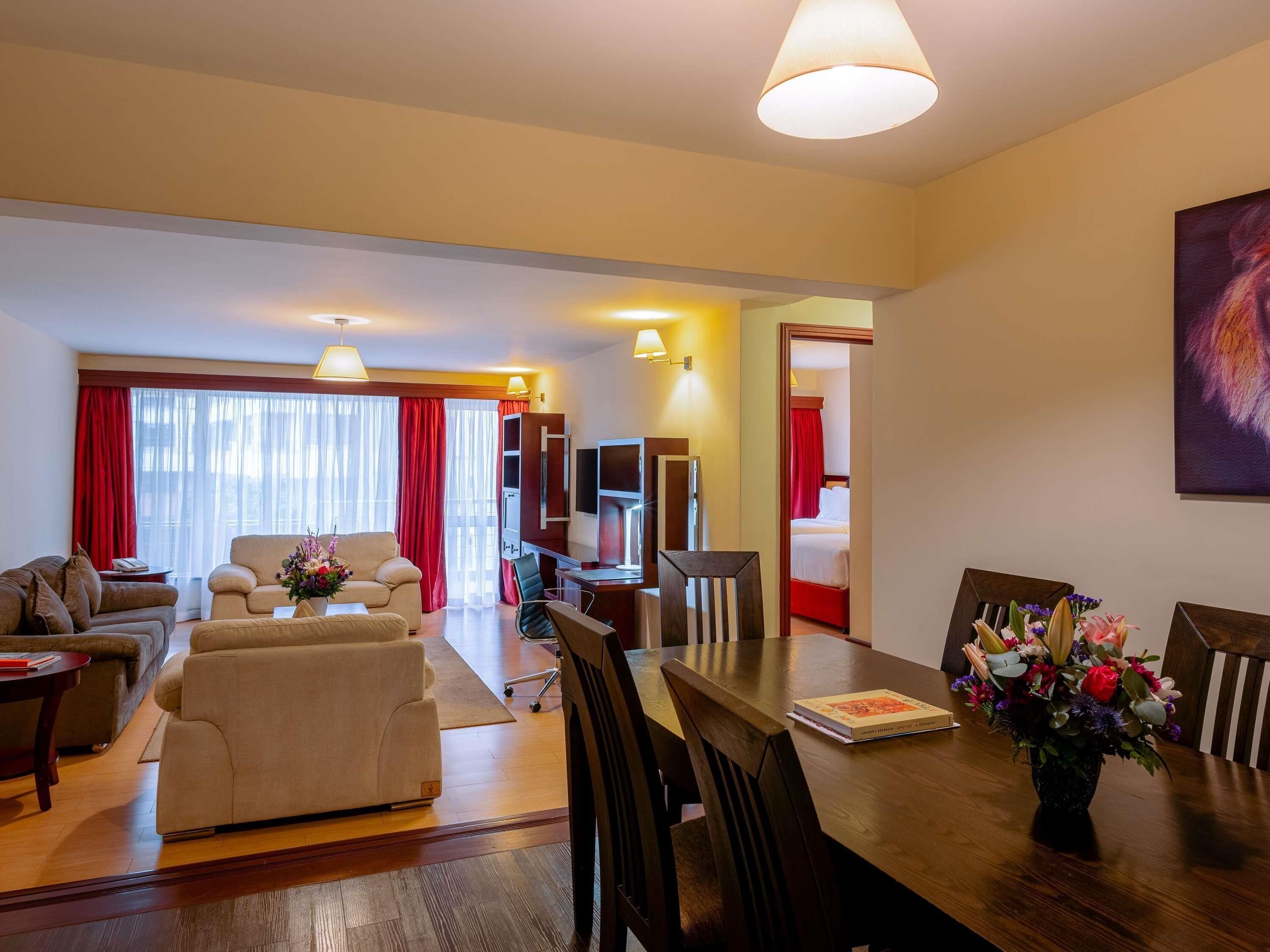 A hotel room with a living area, a dining table with chairs, a TV, and a bed with red curtains.