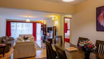 A hotel room with a living area, a dining table with chairs, a TV, and a bed with red curtains.
