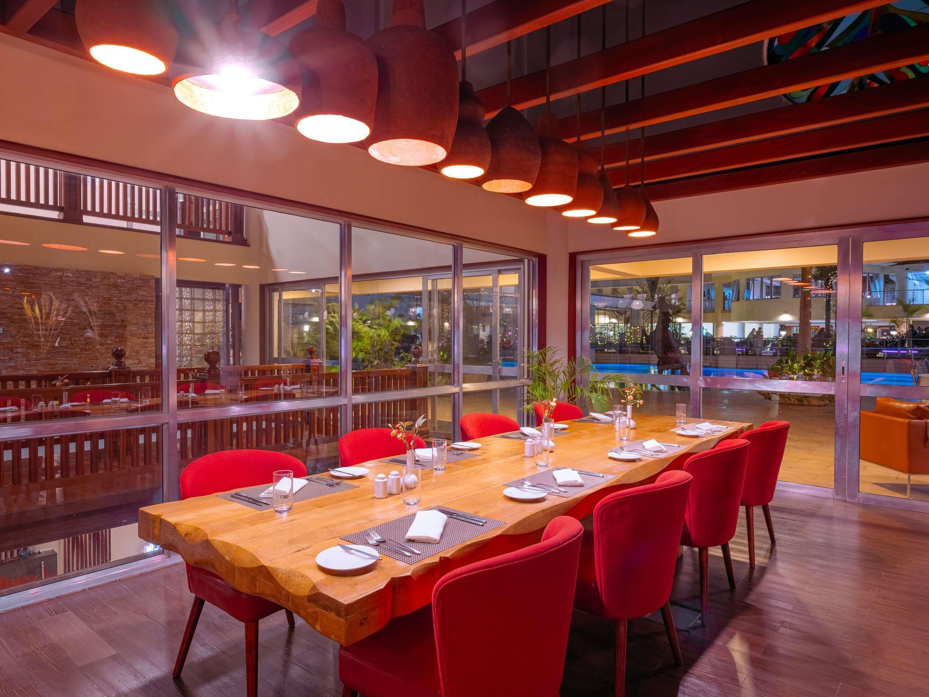 A dining area with a long wooden table, red chairs, and large windows offering views of a pool and outdoor area.