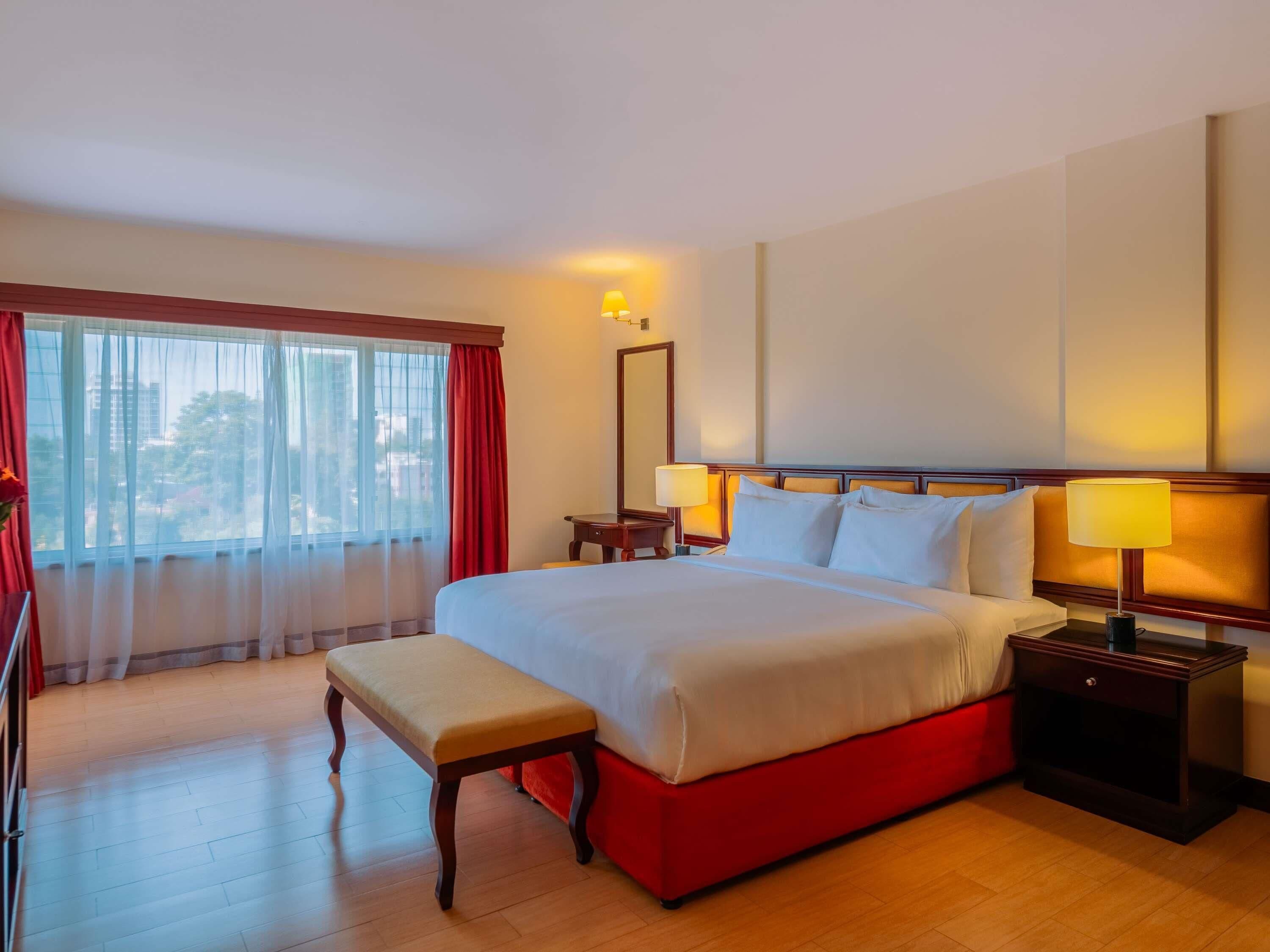A hotel room with a large bed, a bench, two bedside tables with lamps, a mirror, and a window with curtains.