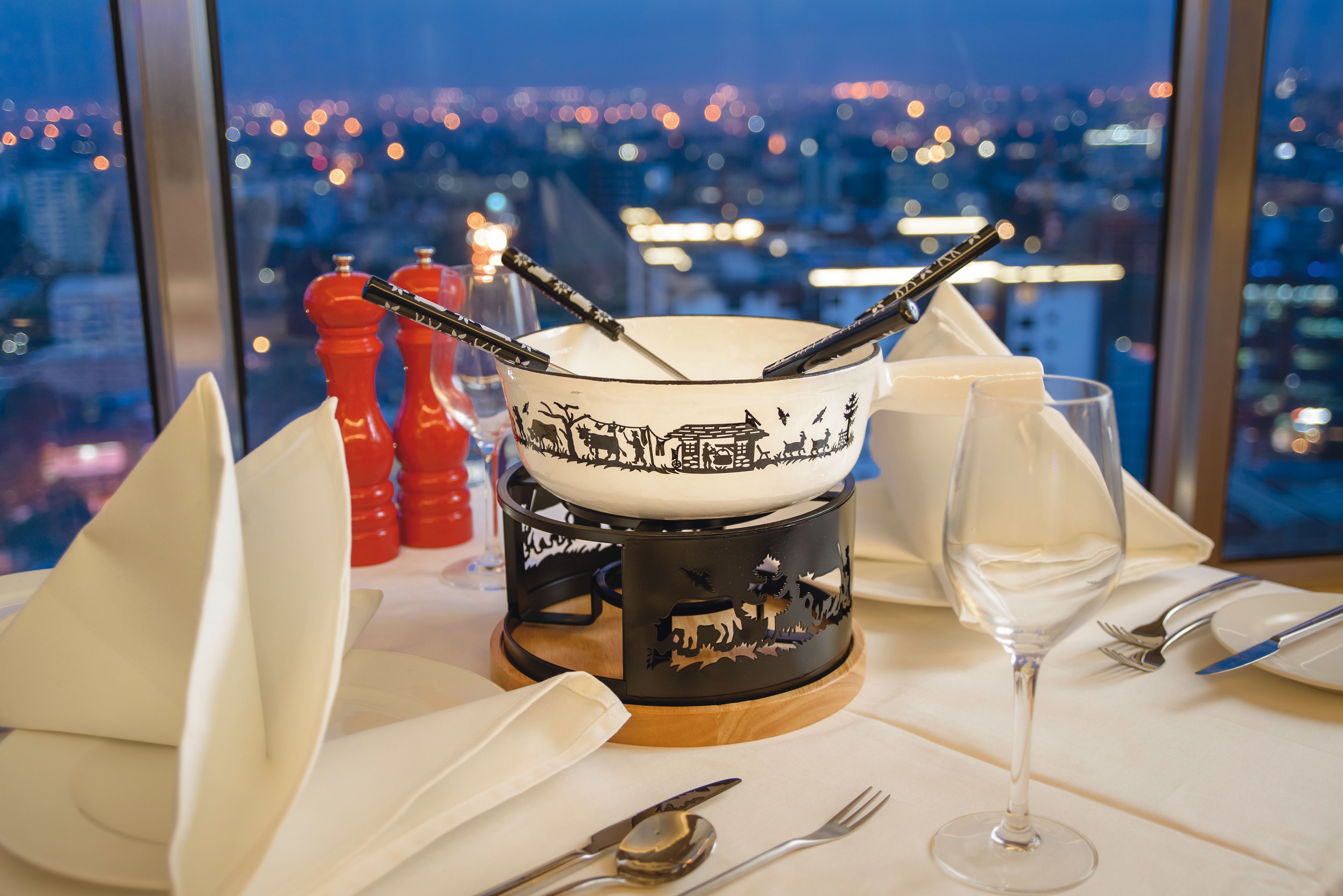 Una mesa de comedor dispuesta con un bol, una copa de vino, cubiertos y servilletas, con una vista de la ciudad al fondo.
