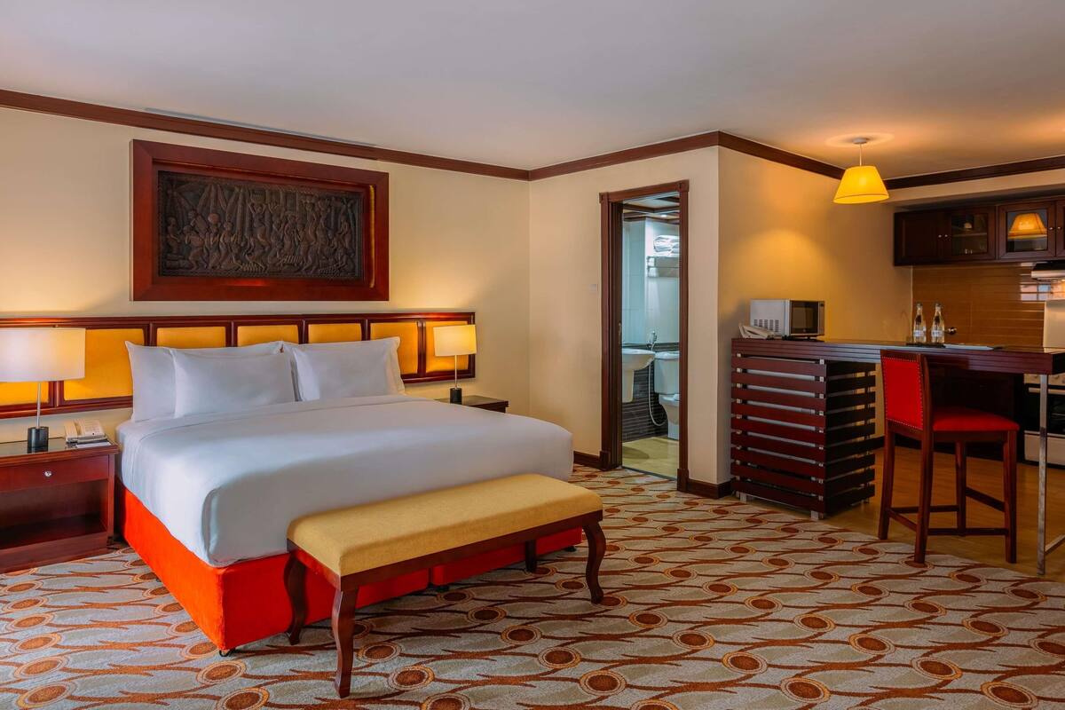 Een hotelkamer met een groot bed, nachtlampjes, een bankje, een bureau met stoel, een kitchenette en een badkamer die door een geopende deur zichtbaar is.