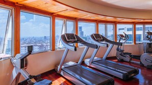 Une salle de sport équipée de tapis de course, d’un rameur et d’un vélo d’appartement, avec de grandes fenêtres donnant sur la ville.