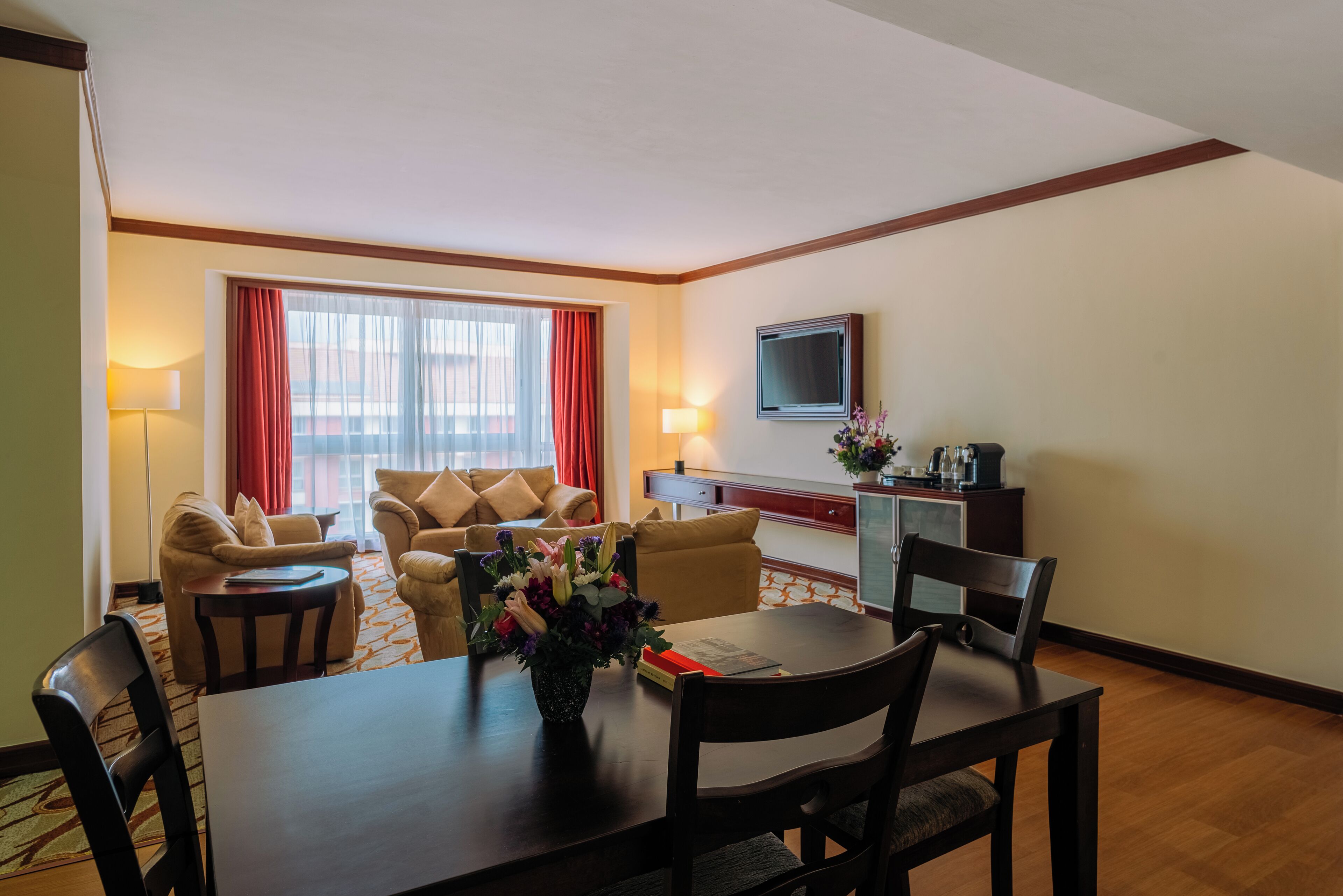 A hotel room with a dining area, a sofa set, a TV, and a desk with a lamp.
