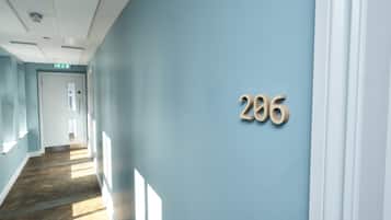 A hallway with a light blue wall and wooden flooring, leading to a door with an emergency exit sign.