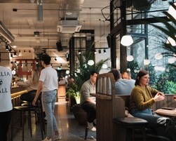 Interior de café moderno com pé-direito alto, tubulação exposta e luz natural proveniente de grandes janelas.