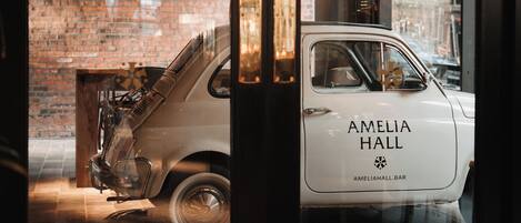 Une voiture ancienne, sur laquelle on peut lire les mots « AMELIA HALL », est garée devant un bâtiment dont la façade est en briques et qui possède une porte en verre.