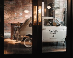 A vintage car with "AMELIA HALL" written on the side parked in front of a building with a brick wall and a glass door.