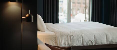 Quarto de hotel com cama, cadeira, abajur e janela com cortinas.