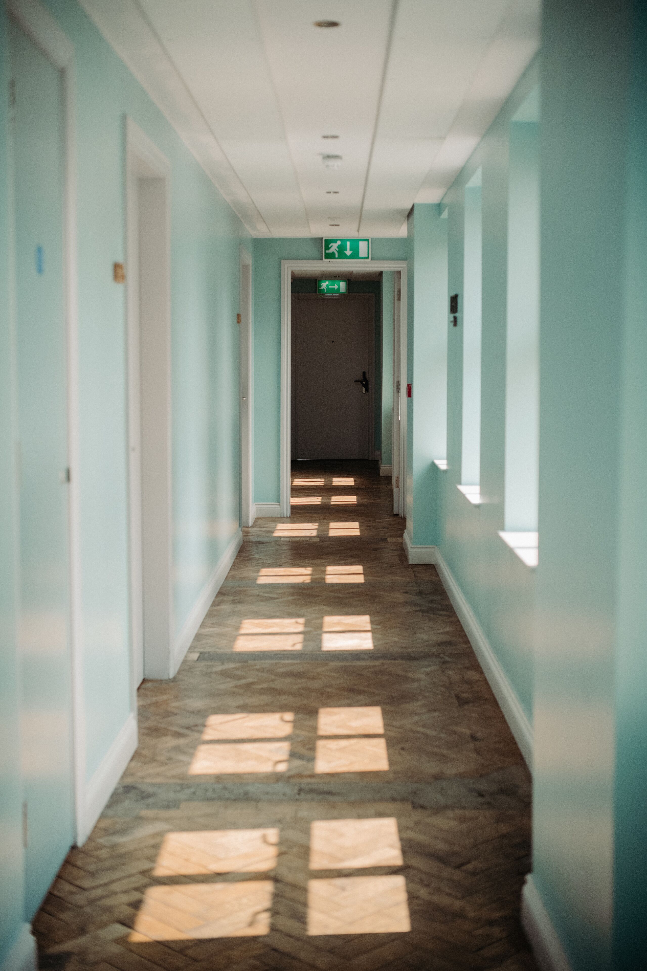 En hall med ljusblå väggar och ett trägolv. Det finns en utrymningsskylt på väggen.