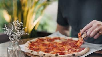 En pizza på ett träbord, tillsammans med en pizzaskärare och en glasvas med torkade blommor.